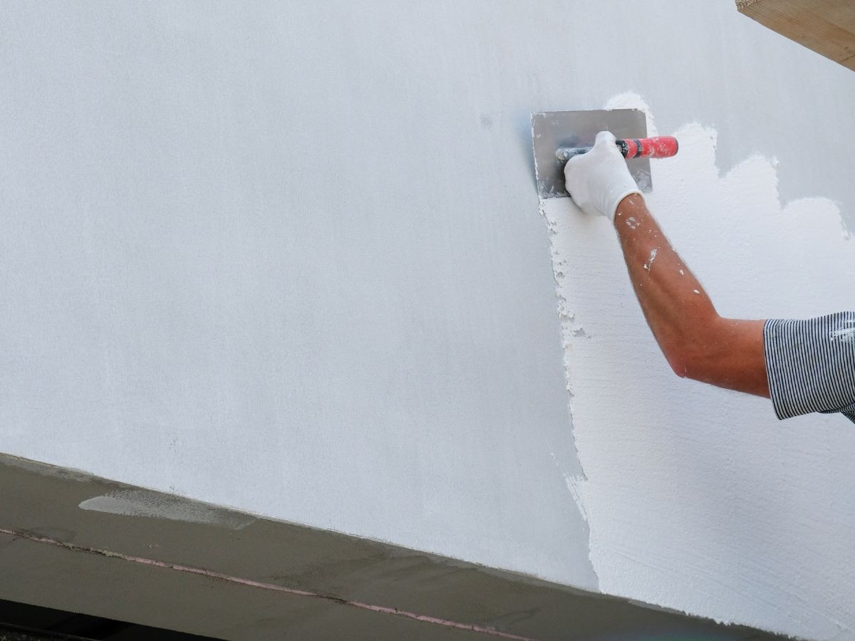 Main gantée talochant un crépi blanc granuleux sur mur gris avec une lisseuse métallique à manche rouge.